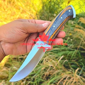 May include: A fixed-blade knife with a polished silver-coloured steel blade and a handle featuring alternating layers of black and orange wood. The handle is secured with three silver-coloured rivets and has a curved pommel at the end.