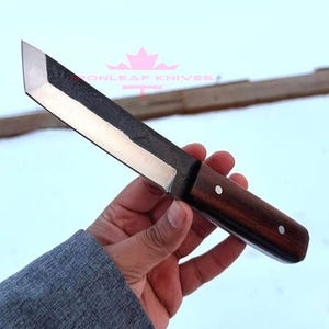 Peut inclure: Un couteau artisanal avec une lame sombre et texturée et un manche en bois poli. La lame a un bord droit et une pointe acérée. Le manche est de couleur marron foncé et est fixé avec deux rivets argentés. Le couteau est tenu dans une main.