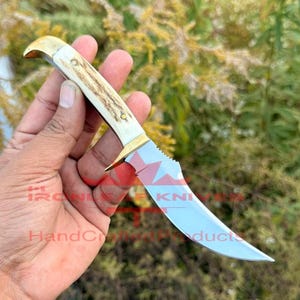 Peut inclure: Un couteau à lame fixe doté d'une lame en acier argenté incurvée, d'une garde en laiton et d'un manche texturé de couleur claire. La lame porte un logo rouge avec le texte IRONLEAF KNIVES et HandCraftedProducts.
