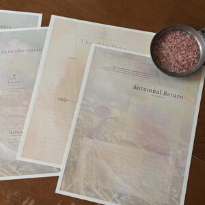 May include: Several printed pages with text and watercolor-style illustrations are arranged on a wooden surface. The pages have titles like "Autumnal Return" and "The Binding." A small metal bowl filled with reddish granules sits nearby.