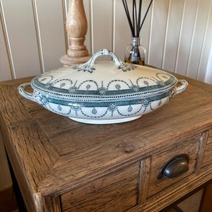 May include: Oval, lidded serving dish with a white base and teal floral and geometric patterns. The dish has two handles and a loop handle on the lid. It sits on a wooden side table with a drawer. A wooden lamp and diffuser are in the background.