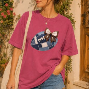 Può includere: Una t-shirt a maniche corte color magenta con una grafica centrale raffigurante un pallone da football americano in patchwork blu, decorato con un fiocco in tessuto marrone e bianco. La maglia ha un girocollo ed è abbinata a una borsa beige.