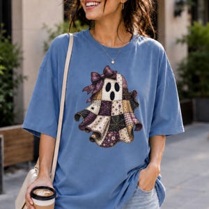 May include: A blue short-sleeve t-shirt featuring a central graphic of a ghost made of various patchwork fabric patterns in shades of brown, purple, and black, accented with a large purple bow on its head and a spider web design.
