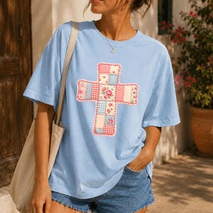 May include: A light blue short-sleeved t-shirt featuring a large central cross graphic composed of a patchwork design. The cross pattern includes various squares with pink, blue, and white floral prints, gingham checks, and polka dot motifs.