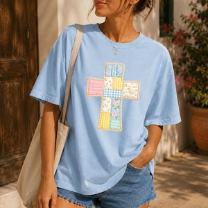 Op de afbeelding: Een lichtblauw t-shirt met korte mouwen en een centraal kruisontwerp bestaande uit diverse pastelkleurige patchworkvierkanten met bloemen-, ruitjes- en effen patronen, inclusief een dunne zilveren ketting en een beige schouderband van een tas.
