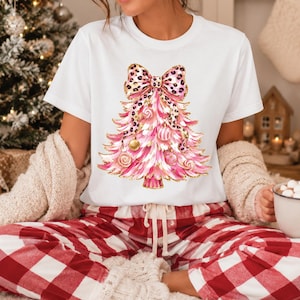 Może przedstawiać: Biały t-shirt z krótkim rękawem z grafiką różowej choinki ozdobionej złotymi bombkami i akcentami przypominającymi cukierki, zwieńczonej dużą kokardą w panterkę. Wzór zawiera złote, brokatowe detale na całej powierzchni choinki.