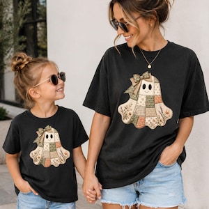 Peut inclure: Deux t-shirts noirs à manches courtes avec un motif central représentant un fantôme composé de divers tissus en patchwork aux tons terreux. Le design du fantôme comprend un petit nœud sur le dessus et des motifs floraux sur le bas.