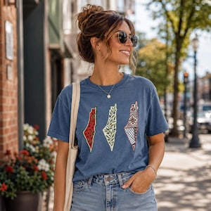 May include: A blue short-sleeved t-shirt featuring three distinct map-shaped graphic designs. The left graphic has a red watermelon pattern, the center graphic displays green olive branches, and the right graphic shows a black and white geometric pattern.