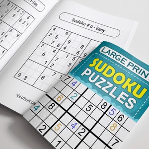 May include: Open book with Sudoku puzzles. The cover reads "LARGE PRINT SUDOKU PUZZLES" in yellow and blue. The pages display partially completed Sudoku grids with numbers. The top page reads "Sudoku # 6 - Easy".