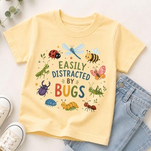 May include: A pale yellow short-sleeve t-shirt featuring a colourful graphic of various insects arranged in a circle around the text "EASILY DISTRACTED BY BUGS". The insects include a ladybird, dragonfly, bee, butterfly, grasshopper, ant, caterpillar, and beetle.