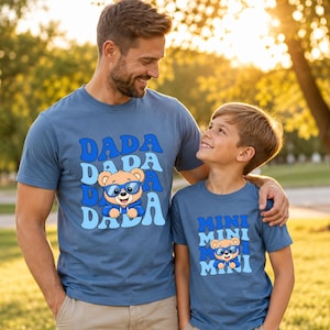 May include: Two blue crew-neck t-shirts featuring a cartoon bear wearing blue sunglasses. The larger shirt displays the text DADA in a repeating wavy blue font, while the smaller shirt displays the text MINI in a matching blue font style.