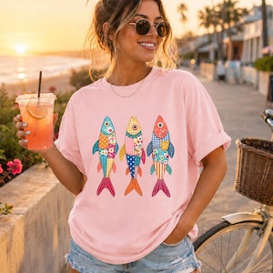 Peut inclure: Un t-shirt à manches courtes rose clair avec une impression graphique représentant trois poissons côte à côte. Chaque poisson est orné d'un motif patchwork coloré composé de divers designs floraux, à pois et à carreaux dans des tons de bleu, jaune, orange et rouge.