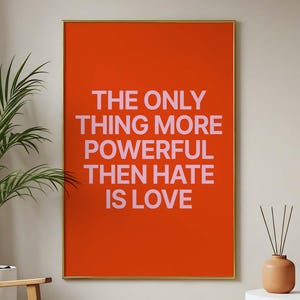 May include: An orange framed art print with the text "THE ONLY THING MORE POWERFUL THEN HATE IS LOVE" in pink. The print is displayed on a beige wall, with a wooden chair, plants, and a white side table in the room.