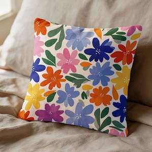 Peut inclure: Coussin carré décoratif avec un motif floral. Le coussin présente un fond crème avec un motif répété de fleurs colorées dans les tons orange, bleu, rose, jaune et violet, avec des feuilles vertes. Le coussin est posé sur un lit.