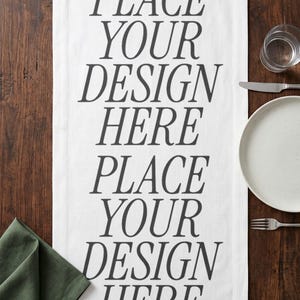 May include: White table runner with the text "PLACE YOUR DESIGN HERE" in a bold, black font. The runner is on a dark wood table, with a plate, silverware, and a glass of water.