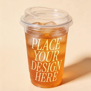Pode incluir: Um copo de plástico transparente com tampa, cheio de chá gelado e cubos de gelo. O texto "PLACE YOUR DESIGN HERE" está impresso em branco no copo. O fundo é de cor pêssego claro.