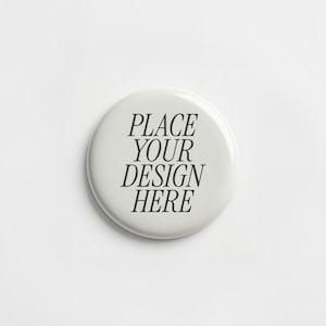 May include: A circular white pin-back button featuring the black text PLACE YOUR DESIGN HERE centered on the surface. The button has a smooth, glossy finish and a rounded edge, presenting a clean and simple design for custom graphic applications.