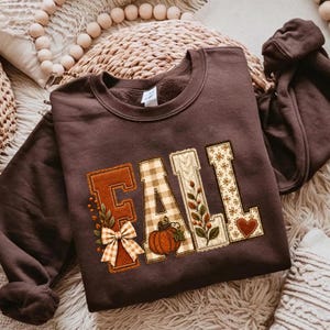 May include: A dark brown crewneck sweatshirt featuring the word FALL in large block letters. The letters are decorated with patterns including a plaid bow, a pumpkin, autumn leaves, and a small heart, all in shades of orange and cream.
