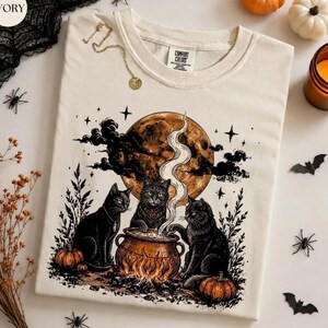 May include: An ivory t-shirt featuring a graphic print of three black cats surrounding a steaming cauldron under a large orange moon. The design includes stars, pumpkins, and foliage. The shirt has a Comfort Colors label at the inner neckline.