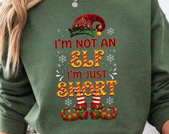 Peut inclure: Sweat-shirt vert avec le texte "I'm not an elf, I'm just short" dans une police festive. Le motif comprend un chapeau et des chaussures d'elfe. La personne porte un pantalon de pyjama à carreaux rouges et blancs et tient une tasse du Père Noël.