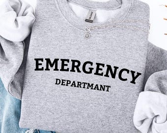 May include: A heather gray sweatshirt with the words "EMERGENCY DEPARTMENT" printed in black. The sweatshirt has a crew neck and long sleeves with white cuffs. The image also includes a pair of blue jeans.