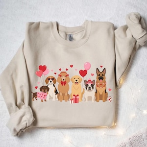 Peut inclure: Sweat-shirt beige avec un motif de sept chiens de dessin animé, des ballons en forme de cœur et des cœurs. Les chiens comprennent un teckel, un beagle, un caniche, un golden retriever, un bouledogue français et un berger allemand. Thème de la Saint-Valentin.