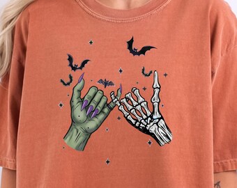May include: Orange-colored t-shirt featuring a Halloween-themed graphic. The graphic depicts two hands, one green and one skeletal, making a pinky promise. Black bats and stars are scattered around the hands.