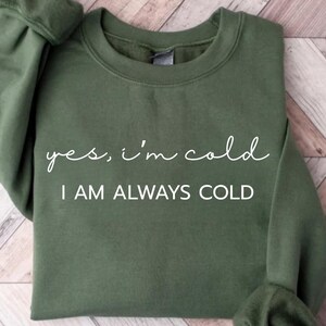 Może przedstawiać: Zielona bluza w kolorze oliwkowym z białym napisem "yes, i'm cold" i "I AM ALWAYS COLD". Bluza z okrągłym dekoltem ma długie rękawy ze ściągaczami. Odzież jest złożona na jasnej powierzchni z usłojeniem drewna.
