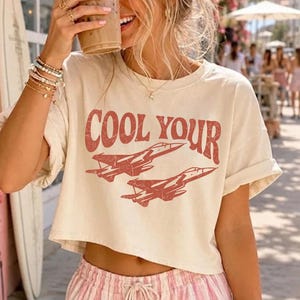 May include: A cream-colored cropped t-shirt featuring the red text COOL YOUR and a graphic illustration of two fighter jets. The shirt has short sleeves and a boxy fit, paired with light pink and white vertical striped drawstring shorts.