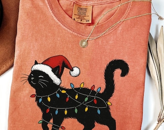 Puede incluir: Una camiseta de color coral con un gato negro que lleva un gorro de Papá Noel y está envuelto en luces navideñas de colores. La camiseta tiene cuello redondo y mangas cortas. Un collar dorado con un colgante está drapeado sobre la camiseta.