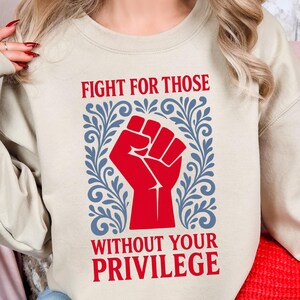 May include: A beige sweatshirt with the text "FIGHT FOR THOSE WITHOUT YOUR PRIVILEGE" in red. The text is above a red fist graphic, framed by blue floral designs. The sweatshirt is a casual style.