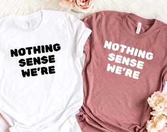 Puede incluir: Dos camisetas, una blanca y otra malva, con el texto "NOTHING SENSE WE'RE" en letras negras y blancas en negrita. Las camisetas se muestran sobre una superficie blanca con detalles florales.