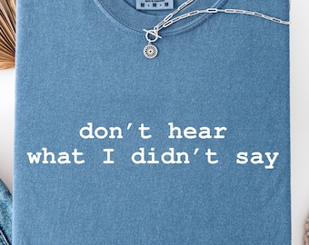 May include: A blue t-shirt with the text "don't hear what I didn't say" in white. A silver necklace with a sun-shaped pendant rests on the shirt. The shirt is folded and has a Comfort Colors tag.