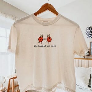 May include: A cream-coloured short-sleeve t-shirt featuring a graphic of two red ladybirds with black spots holding hands. Below the ladybirds, the text reads "the last of the bugs" in a simple black font. The shirt is displayed on a wooden hanger.