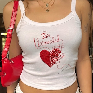 Op de afbeelding: Een wit hemdje met een rood geborduurd ontwerp van een hart dat deels solide is en deels is ontrafeld tot losse draden, met de tekst "I'm Unraveled" erboven en een naald rechtsonder in het ontwerp.