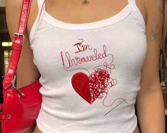 May include: A white tank top featuring a red embroidered graphic of a heart that is partially solid and partially unraveled into loose thread, with the text "I'm Unraveled" above it and a needle at the bottom right of the design.