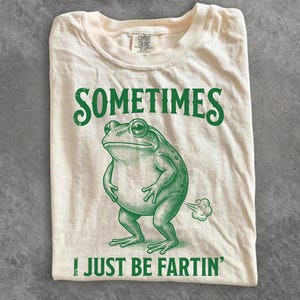 Può includere: T-shirt color crema con grafica verde di una rana e la scritta "SOMETIMES I JUST BE FARTIN'". L'illustrazione della rana è dettagliata, con la scritta "COMFORT COLORS" stampata sull'etichetta.