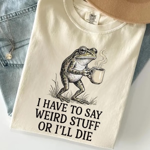 Op de afbeelding: Een crèmekleurig t-shirt met een afbeelding van een kikker die een dampende mok vasthoudt. Onder de kikker staat de tekst: "I HAVE TO SAY WEIRD STUFF OR I'LL DIE". Het t-shirt heeft een ronde hals en een Comfort Colors label in de nek.