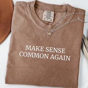 Puede incluir: Una camiseta marrón con el texto "MAKE SENSE COMMON AGAIN" en blanco. La camiseta tiene cuello redondo y mangas cortas. Un collar plateado con un pequeño colgante está sobre la camiseta. Un sombrero marrón es parcialmente visible.