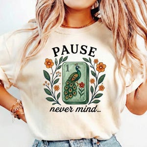 May include: A cream-colored t-shirt featuring a graphic print of a green mahjong tile with a peacock illustration and the number one. The design is framed by orange flowers and green leaves, with the text PAUSE above and never mind below.