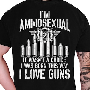 May include: A black t-shirt featuring white distressed text that reads I'M AMMOSEXUAL, IT WASN'T A CHOICE, I WAS BORN THIS WAY, I LOVE GUNS. The design includes a graphic of eight various bullet cartridges arranged in a row with a gear emblem.
