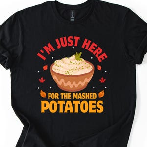 Puede incluir: Camiseta negra con el texto "I'M JUST HERE FOR THE MASHED POTATOES" en rojo y naranja. El diseño incluye un cuenco de puré de patatas y hojas de otoño.