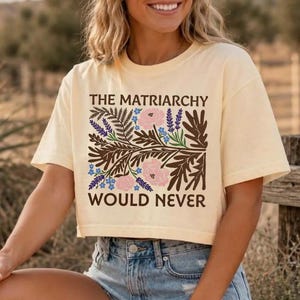 Könnte beinhalten: Ein elfenbeinfarbenes, kurzärmeliges bauchfreies Oberteil mit einem braunen Grafikdruck und dem Text THE MATRIARCHY WOULD NEVER, umgeben von Illustrationen aus braunen Farnblättern, rosa Blüten und kleinen blauen Blumen auf der Vorderseite.