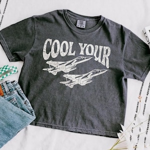 May include: A charcoal gray short-sleeve t-shirt featuring a distressed white graphic of two fighter jets and the text COOL YOUR. The shirt has a crew neckline and a faded, washed-out appearance, with a visible brand tag inside the collar.