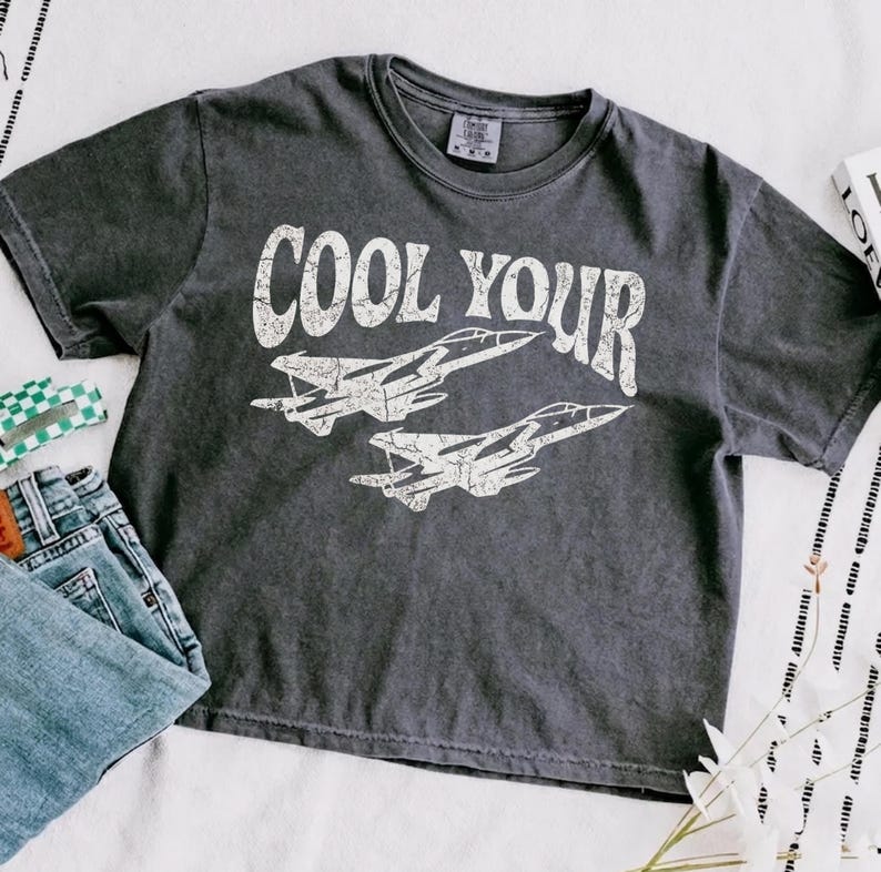 May include: A charcoal gray short-sleeve t-shirt featuring a distressed white graphic of two fighter jets and the text COOL YOUR. The shirt has a crew neckline and a faded, washed-out appearance, with a visible brand tag inside the collar.
