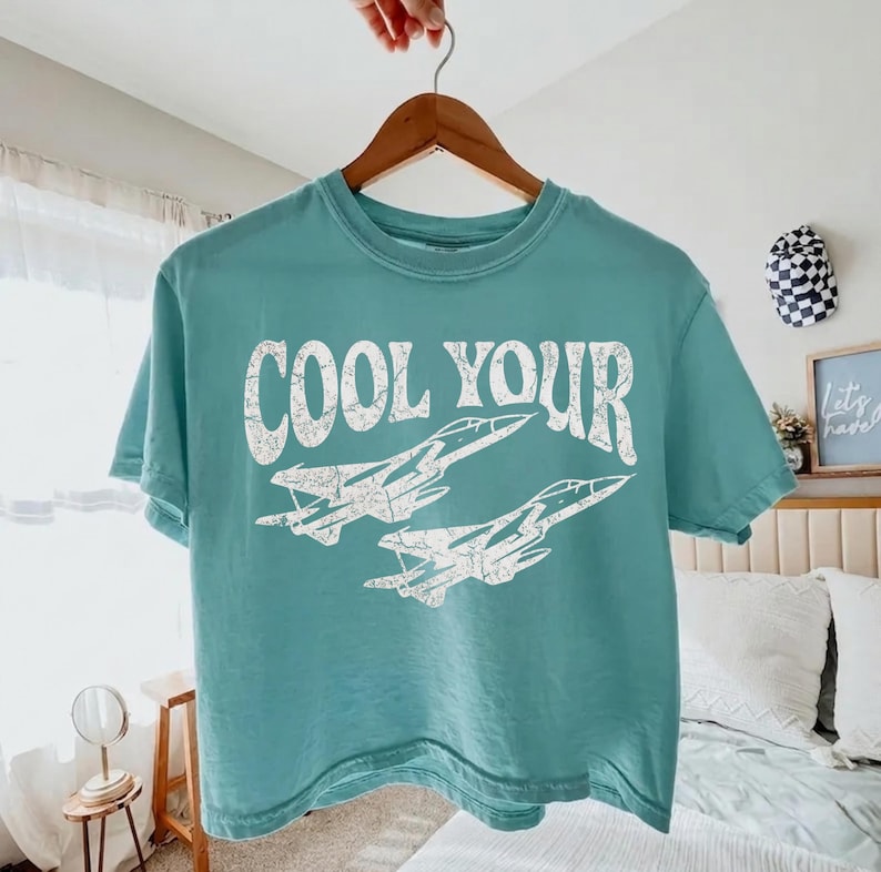 May include: A teal short-sleeve t-shirt featuring the white distressed text "COOL YOUR" above a graphic illustration of two fighter jets flying in formation. The shirt is displayed on a wooden hanger against a neutral indoor setting.