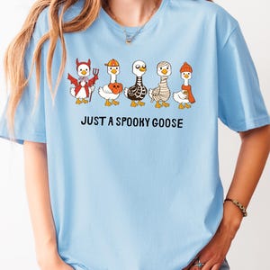 May include: Light blue t-shirt featuring a graphic of five cartoon geese dressed in Halloween costumes. The geese are wearing a devil costume, a pumpkin hat, a skeleton costume, a mummy costume, and a winter hat. The text "JUST A SPOOKY GOOSE" is printed below the geese.