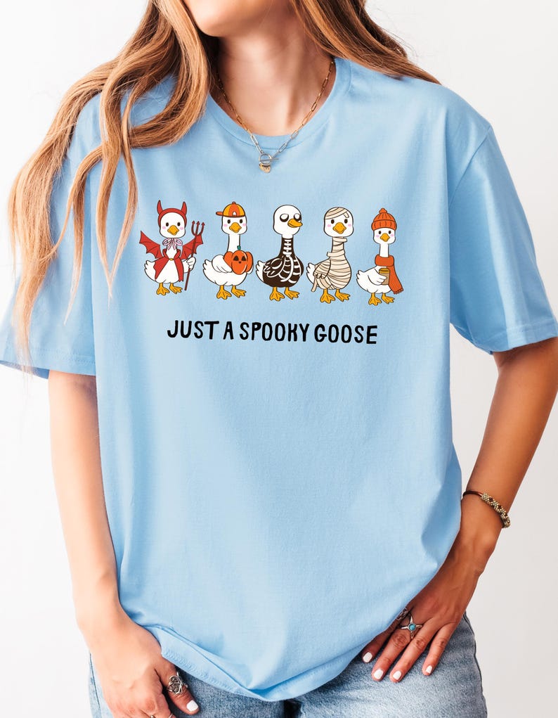 May include: Light blue t-shirt featuring a graphic of five cartoon geese dressed in Halloween costumes. The geese are wearing a devil costume, a pumpkin hat, a skeleton costume, a mummy costume, and a winter hat. The text "JUST A SPOOKY GOOSE" is printed below the geese.