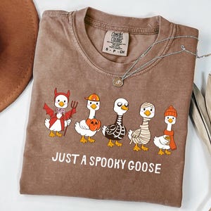 May include: Brown t-shirt with the text "JUST A SPOOKY GOOSE". The shirt features a graphic of five geese dressed in Halloween costumes: a devil, a pumpkin, a skeleton, a mummy, and a goose with a coffee cup.