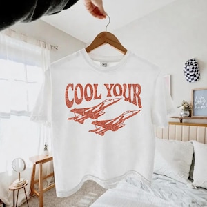 May include: A white short-sleeve t-shirt featuring a distressed red graphic of two fighter jets flying in formation. Above the jets, the text reads COOL YOUR in a bold, retro-style font. The shirt is displayed on a wooden hanger.